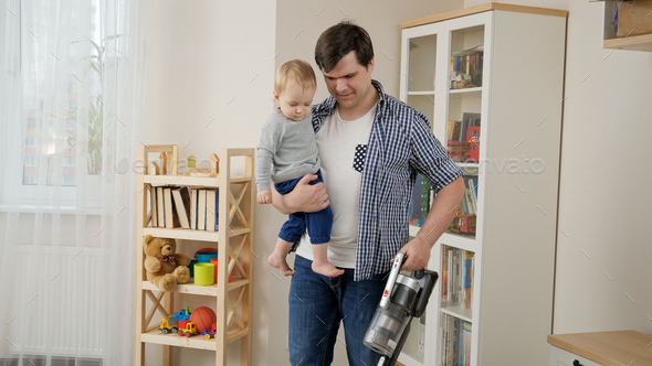 Young father holding his baby son while doing house cleanup and using vacuum cleaner. - Stock Photo - Images