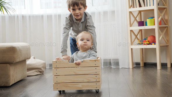 Older boy riding his baby brother in wooden toy cart in living room. - Stock Photo - Images