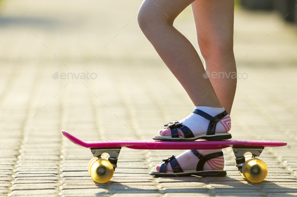 Child slim legs in white socks and black sandals on plastic pink skateboard