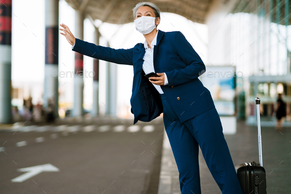 Grey asian woman in face mask waving for taxi on airport parking