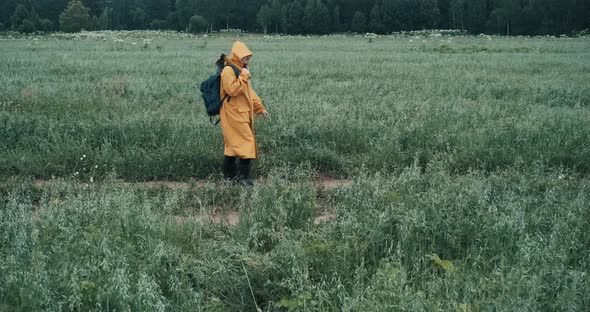 Man in Raincoat Carrying an Axe and Backpack is Walking Through Field
