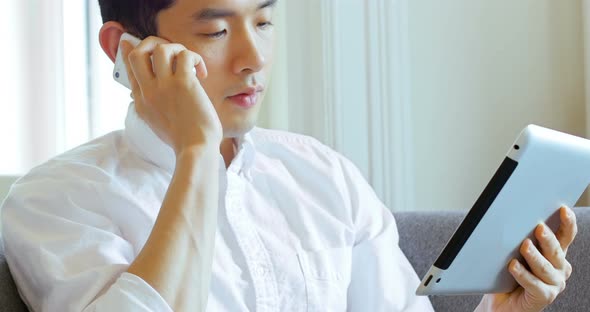 Man talking on mobile phone while using digital tablet in living room 4k alt