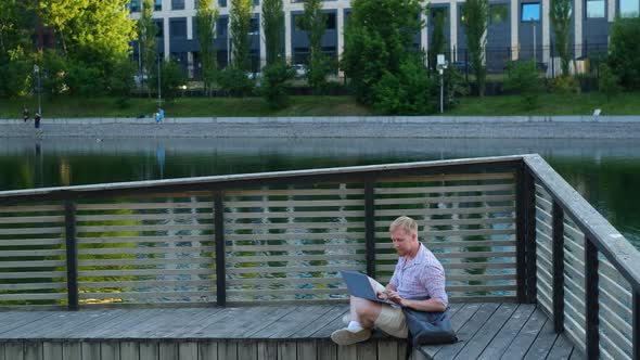 male freelancer working with laptop in the city park by the lake