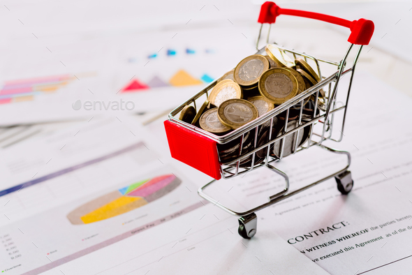 Mini supermarket cart full of coins with data chart and profits in commerce.