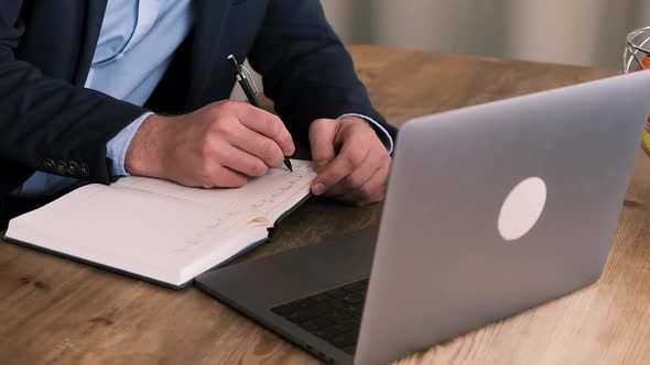 Side View of a Business Man in a Suit Filling Out a Planner or Notepad Next to an Open Laptop on a