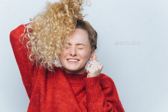 Woman feels excited, keeps eyes shut and clenches fists, has curly light pony tail.