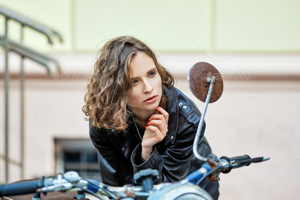 Pretty lady in leather coat and wild curly hair looking to rer view mirror of old vintage motorbike