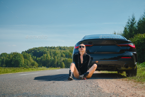 Strong woman in bodysuit and rough boots sits on asphalt by her powerful racing car - Stock Photo - Images