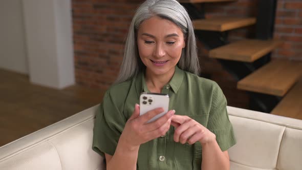 Happy Asian Middleaged Woman Using Smartphone Sitting on the Couch at Home alt