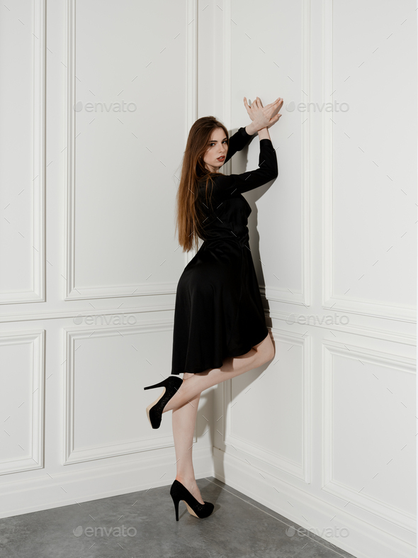 Fashion model stands in the corner of the room, leaning to the wall and lifting one leg
