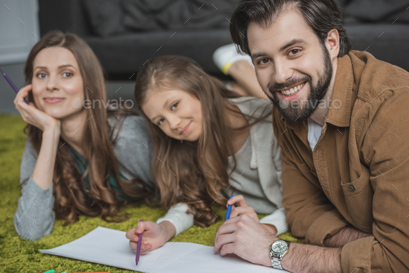 happy parents and daughter drawing with felt-tip pens on floor and looking at camera