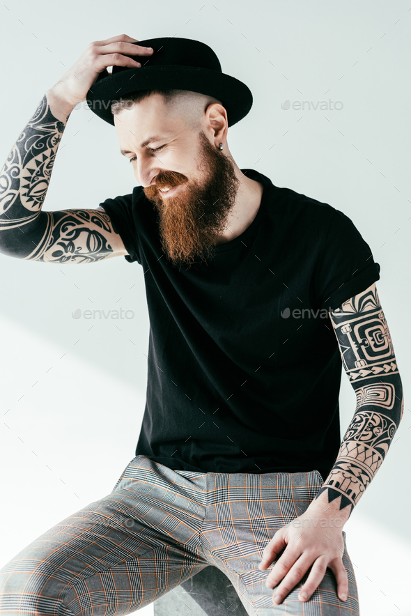 smiling bearded tattooed man touching hat on white Stock Photo by  LightFieldStudios