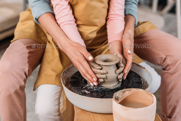 cropped view of teacher and kid making ceramic pot together on pottery wheel