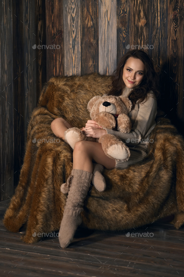 Cute girl in knee socks and sweater with Teddy bear in her hands sitting in armchair in wooden room.
