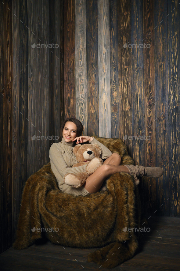 Cute girl in knee socks and sweater with Teddy bear in her hands sitting in armchair in wooden room.