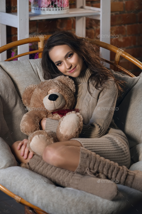 Cute girl in knee socks and sweater with Teddy bear in her hands sitting in armchair in fancy room