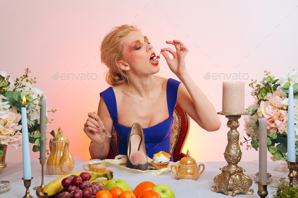 Pretty lady with colorful makeup sitting behind table and eating cherry as a dessert