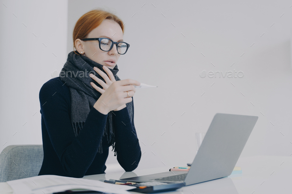 Young white woman is sick while working in office in scarf and looking at thermometer.