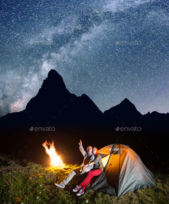 Couple hikers in camp at night near campfire and tent against mountains background and starry sky.