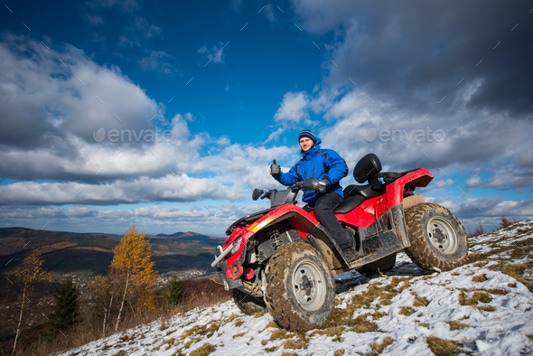 Man with red quad bike on snowy mountain top under the blue cloudy sky.