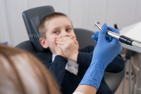 Kid frightened by dentists covers his mouth and dentist in gloves holding in hand dental drill bit