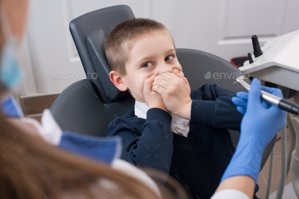 Kid frightened by dentists covers his mouth and dentist in gloves holding in hand dental drill bit