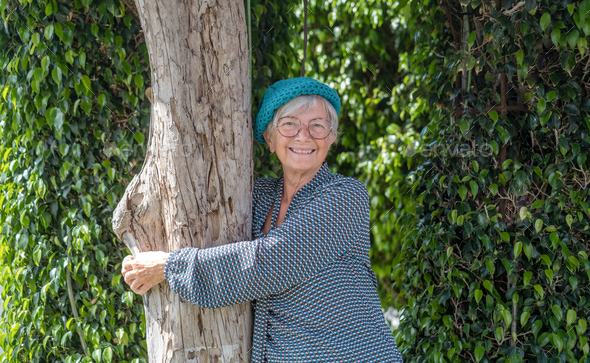 Smiling senior woman in the garden hugging a tree trunk looking at camera, earth day concept