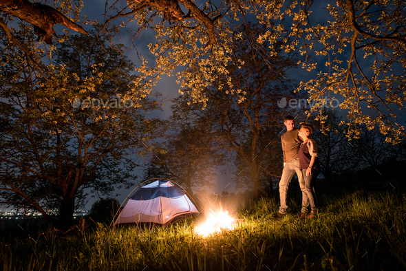 Tourists at a campfire near tent under trees and beautiful night sky full of stars. Night camping