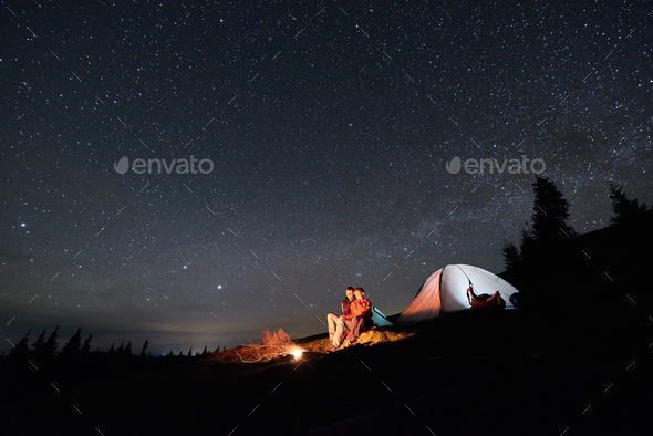 Tourists near campfire and tent under night starry sky