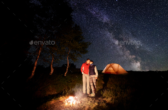 Loving couple enjoying each other standing by fire at night under evening starry sky with Milky way