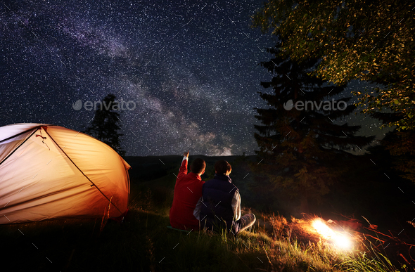 Back view male shows female up on evening starry sky near tent and bonfire on background mountains