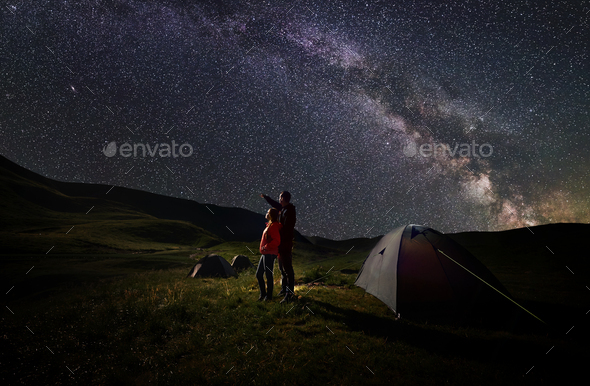 Guy and girl stand near tents under starry sky against backdrop of mountains covered by greenery