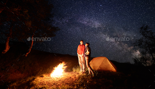 Young people look at each other standing near fire during night camping under starry sky