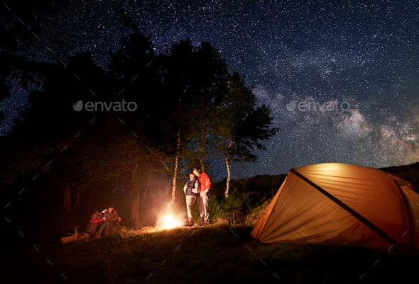 Two young couples at campfire under bright starry sky near tent on background of trees