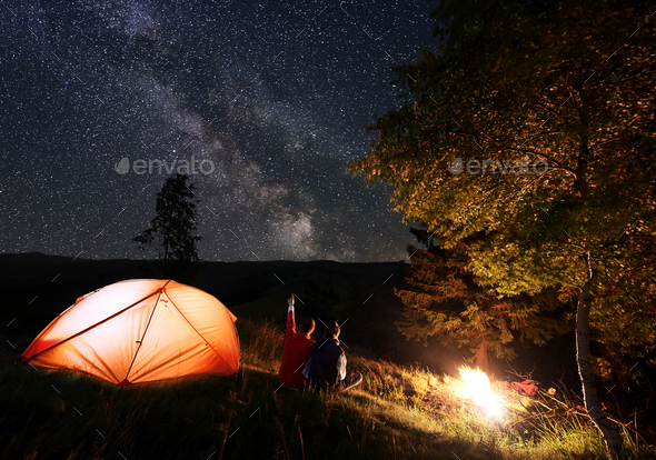 Man shows woman up on evening starry sky at Milky way near tent and bonfire on background mountains