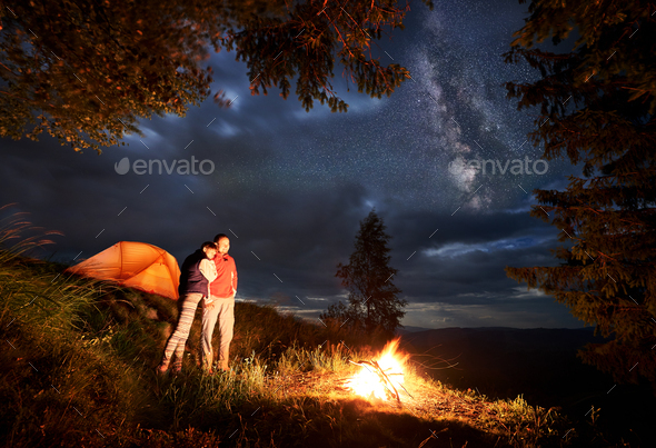 Romantic evening of young couple in mountains by fire under the starry sky
