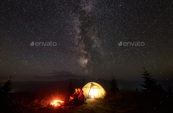 Young couple hikers resting near illuminated tent, camping in mountains at night under starry sky