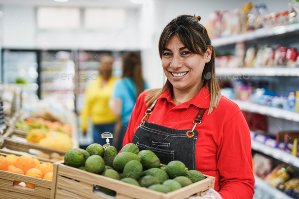 Happy latin woman working inside supermarket while holding wood box with avocados
