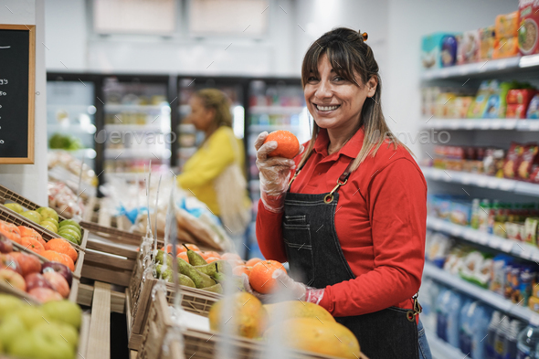 Happy latin woman smiling on camera holding fresh organic fruit while working inside supermarket