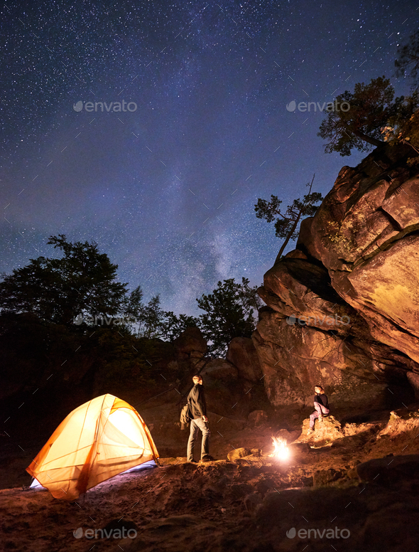 Tourist couple, standing at tent man and girl sitting on big boulder under dark starry sky