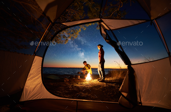Couple man and woman having rest at tourist tent and burning campfire on sea shore near forest