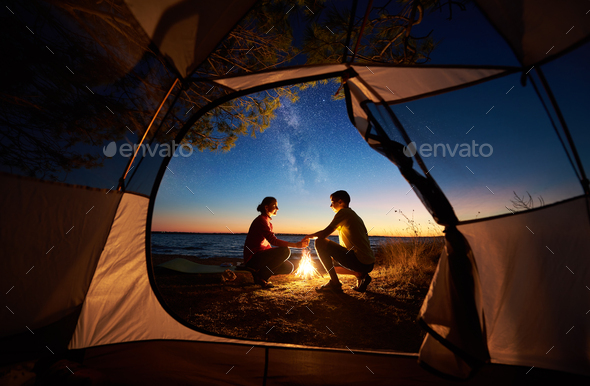 Couple man and woman having rest at tourist tent and burning campfire on sea shore near forest