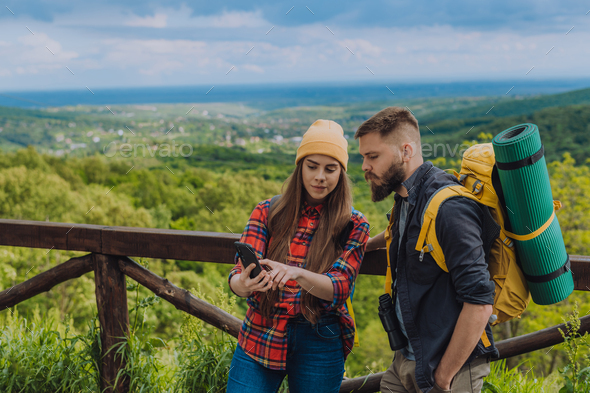 Young couple of hikers using a smartphone while standing on a lookout with camping gear