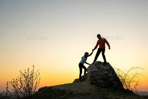 Man and woman hikers helping each other to climb a big stone at sunset in mountains