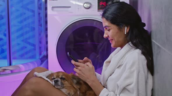 Mixedraces Girl Sitting on Floor with Dog and Watching Video on Smartphone While Washing Machines