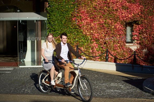 Young couple, handsome man and blond woman cycling tandem bike by building overgrown with red ivy.