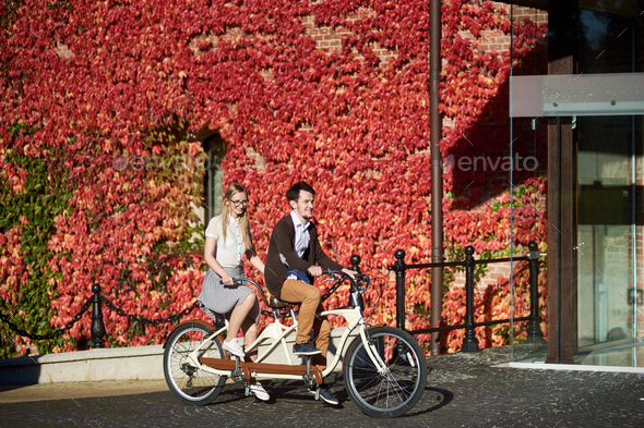 Young couple, handsome man and blond woman cycling tandem bike by building overgrown with red ivy.