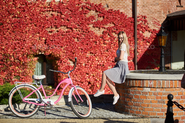 Attractive blond girl at pink lady bicycle on sunny day on wall overgrown with red ivy background.