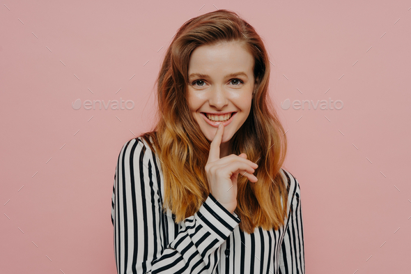 Pretty smiling young girl looking at camera touching lips while standing against pink wall