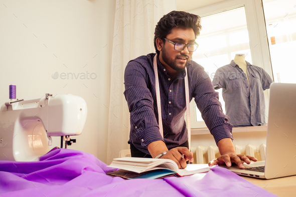handsome indian tailor man In a stylish shirt workinh with violet cotton textile at home
